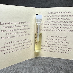 May include: A small glass bottle of perfume with a white label and gold lettering. The bottle is open and the label reads "Eau de Parfum Annick Goutal." The bottle is sitting on a white card with text in French and English describing the perfume.