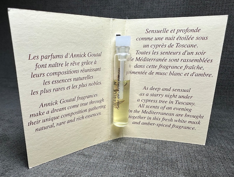 May include: A small glass bottle of perfume with a white label and gold lettering. The bottle is open and the label reads "Eau de Parfum Annick Goutal." The bottle is sitting on a white card with text in French and English describing the perfume.