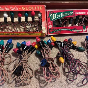 Op de afbeelding: Twee vintage kartonnen dozen met het label Christmas Tree Lights by Glolite en Worthmore Indoor Lights, elk met een set van 7 lampjes. Hieronder liggen vijf verwarde strengen met veelkleurige gloeilampen met zwarte fittingen en bruine gedraaide draden.