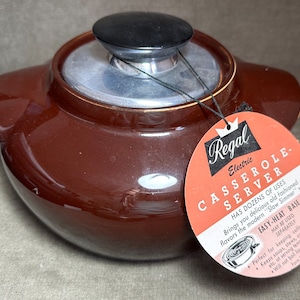 May include: Brown ceramic casserole dish with a silver lid and a black knob. The dish is labeled "Regal Electric Casserole-Server" and has a tag that reads "Has dozens of uses. Brings you delicious old fashioned flavors the modern "Slow Simmer" way. Easy-Heat Base. May be used separately. Perfect for keeping coffee hot. Keeps soups, stews, casseroles, etc. at serving temperatures. Will not boil liquids. Form 97."