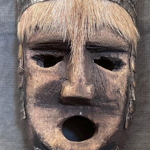 May include: A carved wooden mask with a weathered appearance. The mask features a prominent nose, open mouth, and eye openings. It has a fur-like fringe and a decorative band across the forehead. The mask's overall design suggests a tribal or ceremonial origin.