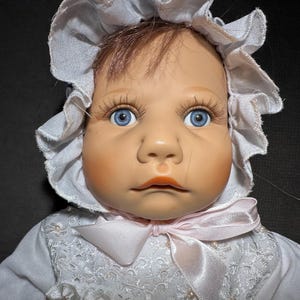 May include: A porcelain baby doll with blue eyes and long eyelashes. The doll wears a white bonnet and a white dress with lace and a pink ribbon. The doll's hair is brown.