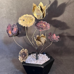 May include: A metal flower sculpture with various flower shapes and colors, including gold, purple, and bronze. The flowers are arranged in a black, geometric pot filled with white pebbles. The sculpture is a decorative item.