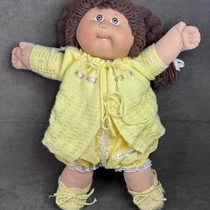 Könnte beinhalten: Vintage Cabbage Patch Kid Puppe mit braunen Garnhaaren und einem lächelnden Gesicht. Die Puppe trägt einen gelben Strickcardigan, passende Shorts mit Vichy-Muster und gelbe Strickschuhe. Das Outfit ist mit weißer Spitze verziert.