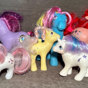 May include: A collection of vintage toy ponies in various colors, including pink, purple, blue, and yellow. Each pony has a flowing mane and tail, with some featuring star and moon designs. The ponies are standing on a neutral surface.