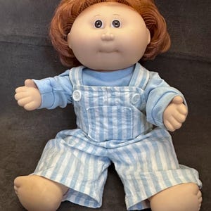 May include: A vintage doll with a round face, brown eyes, and auburn hair. The doll is wearing a light blue long-sleeved shirt and blue and white striped overalls. The doll is sitting with its arms and legs outstretched.