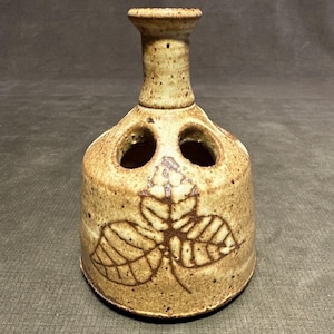 May include: A speckled, tan ceramic toothbrush holder with a flared top and three holes. The cylindrical body features a brown leaf design. The holder is a decorative bathroom accessory.