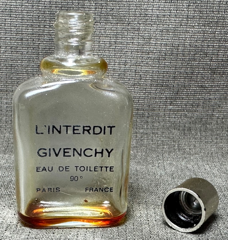 May include: A clear glass bottle of L'Interdit perfume by Givenchy. The bottle has a black label with the text "L'Interdit Givenchy Eau de Toilette 90&deg; Paris France". The bottle has a silver cap.
