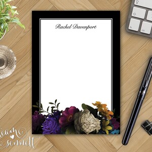 May include: A personalized notepad with a black border and floral design. The notepad features the name "Rachel Davenport" at the top and a colorful arrangement of flowers at the bottom, including purple, white, and yellow blooms. A black pen and paper clip are on the wooden surface.