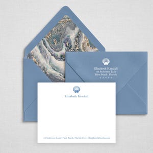 May include: A white stationery card and two dusty blue envelopes. The card features a blue seashell icon, the name Elizabeth Kendall, and address text. One envelope is open, revealing a marbled blue and gold liner, while the other is closed.