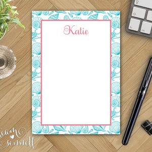 May include: A white notepad with a pink border and a blue seashell pattern. The name "Katie" is written in pink at the top of the notepad.