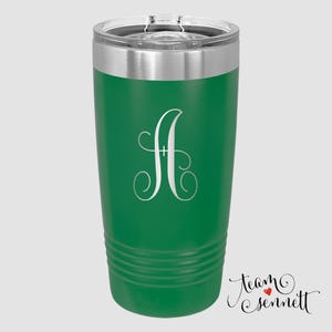 May include: A green insulated tumbler with a silver rim and a clear lid. The tumbler features a large, elegant, silver letter "A" in a script font. The bottom of the tumbler has horizontal grooves. The words "Team Sennett" are written in cursive.