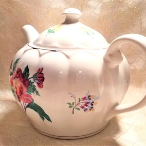 May include: White teapot with a floral design featuring pink, red, and green flowers. The teapot has a rounded body and a curved handle.