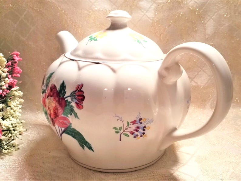 May include: White teapot with a floral design featuring pink, red, and green flowers. The teapot has a rounded body and a curved handle.