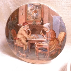 May include: A vintage ceramic tile depicting a scene of two people playing cards at a table. The tile is painted in a realistic style with muted colors and features a dog lying on the floor. The tile is signed "A. Blair".