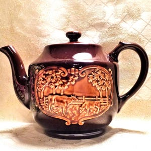 May include: A brown ceramic teapot with a raised design of cows in a field. The teapot has a heart-shaped design around the cows.