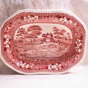 May include: A pink and white oval serving platter with a raised edge and a floral design. The center of the platter features a pink and white landscape scene with a bridge and a building.