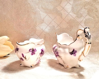 May include: A vintage porcelain sugar and creamer set with a delicate floral design. The set features a white base with purple flowers and gold accents. The creamer has a unique handle shaped like a bird's head.