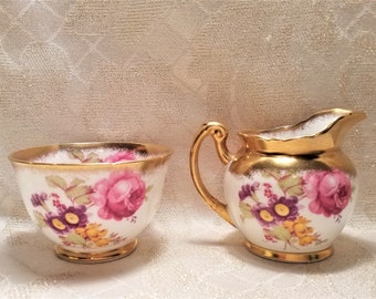 May include: A white creamer and sugar bowl set with a floral design and gold accents. The floral design features pink, purple, and yellow flowers.