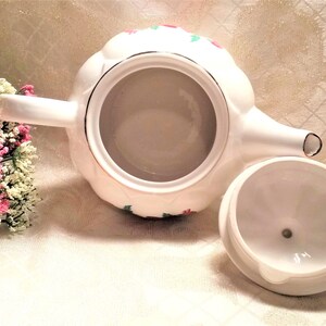 May include: White teapot with a floral design in red, green, and gold. The teapot has a lid and a spout. The teapot is sitting on a white surface.