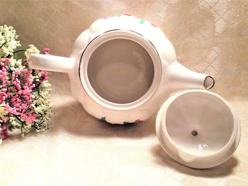 May include: White teapot with a floral design in red, green, and gold. The teapot has a lid and a spout. The teapot is sitting on a white surface.