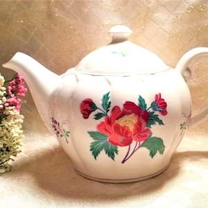 May include: White teapot with a floral design featuring red, green, and yellow flowers. The teapot has a lid and a handle.