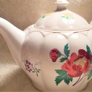 May include: White teapot with a floral design. The teapot has a lid with a gold trim and a floral design. The teapot has a round body with a floral design. The teapot has a spout and a handle.