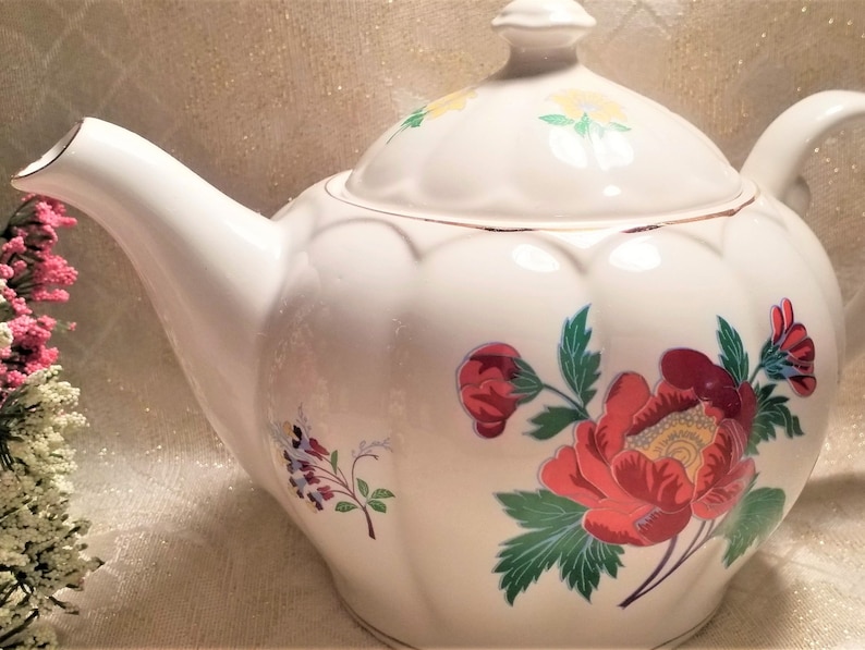 May include: White teapot with a floral design. The teapot has a lid with a gold trim and a floral design. The teapot has a round body with a floral design. The teapot has a spout and a handle.