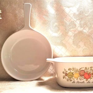May include: A white ceramic baking dish with a floral design and the text "Le Persil La Sauge" on the front. The dish is next to a white ceramic skillet with a long handle.