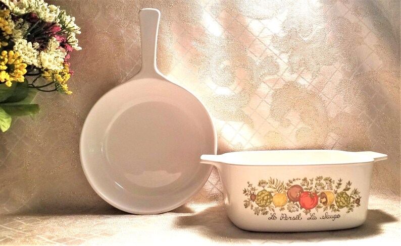 May include: A white ceramic baking dish with a floral design and the text "Le Persil La Sauge" on the front. The dish is next to a white ceramic skillet with a long handle.