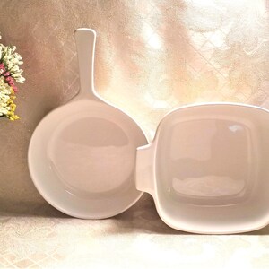 May include: Two white ceramic baking dishes, one round with a handle and one square. Both dishes are empty and have a glossy finish.