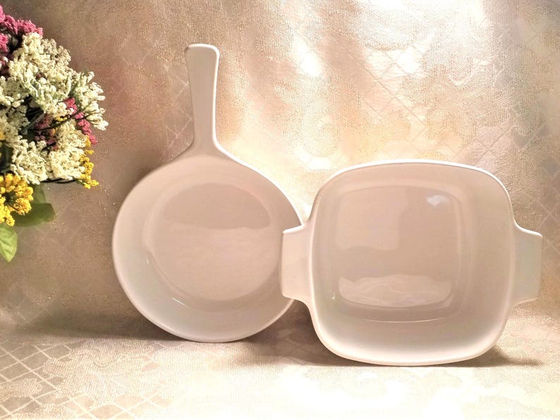 May include: Two white ceramic baking dishes, one round with a handle and one square. Both dishes are empty and have a glossy finish.