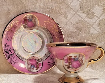 May include: A pink and gold teacup and saucer set with a floral design and a couple in a romantic scene. The saucer has a pearlescent finish.