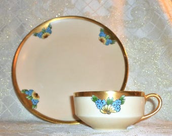 May include: A vintage teacup and saucer set with a cream-colored background and gold trim. The set features a floral design with blue and yellow flowers.