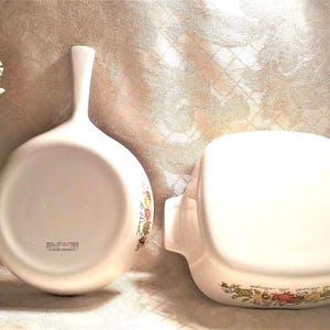 May include: Two white ceramic cookware pieces with floral designs. One is a round skillet with a handle and the other is a square baking dish.