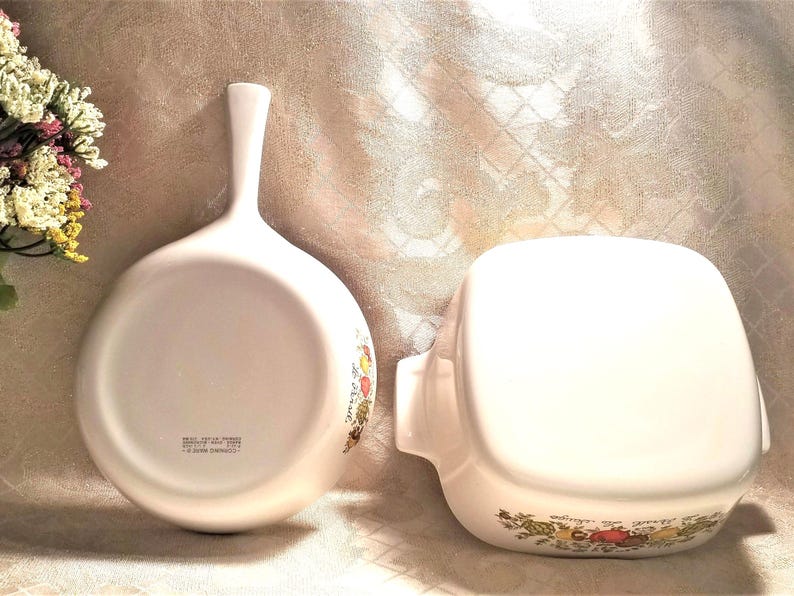 May include: Two white ceramic cookware pieces with floral designs. One is a round skillet with a handle and the other is a square baking dish.