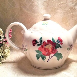 May include: White teapot with a floral design featuring red, yellow, and green flowers. The teapot has a rounded body and a spout on the right side.