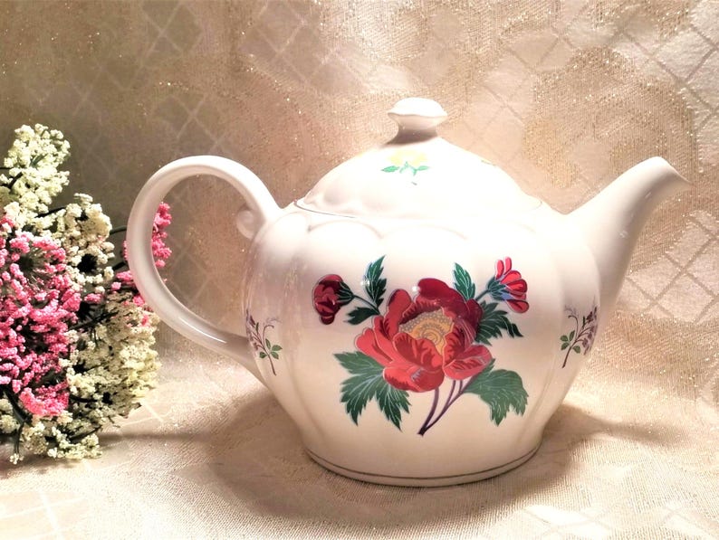 May include: White teapot with a floral design featuring red, yellow, and green flowers. The teapot has a rounded body and a spout on the right side.