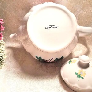 May include: White teapot with a floral design and a matching lid. The teapot has a handle and spout. The lid has a knob on top. The teapot is decorated with a floral pattern in shades of green, yellow, and red. The teapot has the text "Perfume Laura Ashley London Paris New York" on the bottom.