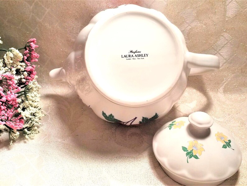 May include: White teapot with a floral design and a matching lid. The teapot has a handle and spout. The lid has a knob on top. The teapot is decorated with a floral pattern in shades of green, yellow, and red. The teapot has the text "Perfume Laura Ashley London Paris New York" on the bottom.