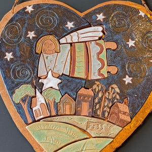 May include: Heart-shaped ceramic wall hanging with a celestial design. Features an angel holding a star above a village scene. The background is dark blue with white stars and swirls. The border is a metallic gold color.