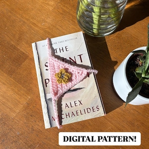 May include: A pink crocheted bookmark with a yellow flower design. The bookmark is on top of a book titled "The Silent Patient" by Alex Michaelides. The text "Digital Pattern!" is at the bottom of the image.
