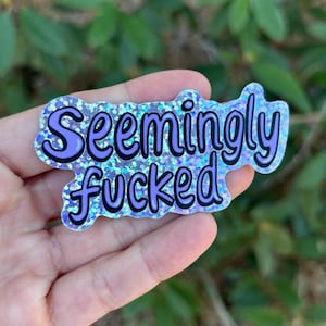 May include: A glittery sticker with the words "Seemingly fucked" in black outlined in purple. The sticker has a light blue and purple glitter background and is held in a hand.