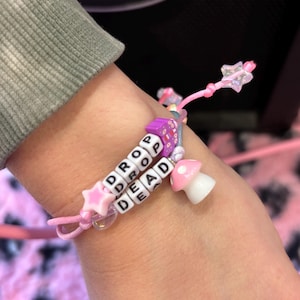 Puede incluir: Pulsera de cuentas rosas con las palabras "DROP DEAD" deletreadas en cuentas de letras blancas. La pulsera presenta dijes de estrella y hongo. La pulsera está en la muñeca de una persona.