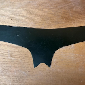 Puede incluir: Una forma negra recortada que se asemeja al contorno de la ropa interior. La forma tiene un borde superior curvo y un borde inferior puntiagudo, con un acabado liso y mate. El objeto está colocado sobre una superficie de madera de color marrón claro.