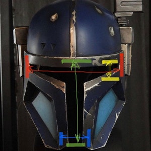 May include: A blue and gray helmet with a visor and a weathered look. The helmet has a rectangular panel on the top and a small rectangular panel on the side. The helmet has a green, yellow, and red stripe across the top.