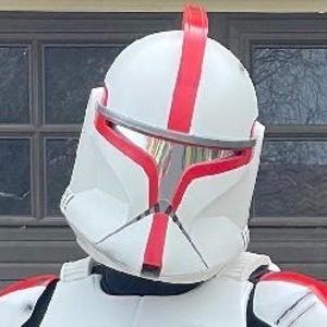 Puede incluir: Un casco blanco y rojo con visera, que recuerda a un soldado clon. El casco presenta detalles rojos en la parte superior y la cara, con una visera plateada. El dise&ntilde;o es detallado y parece ser un disfraz o art&iacute;culo de colecci&oacute;n.