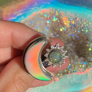 May include: A silver ring featuring a crescent moon and a sun design. The moon has an iridescent, colorful surface, and the sun has a central opal-like stone. The ring is set against a shimmering, rainbow-colored background.