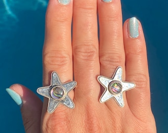 May include: Two silver rings with a star shape and a round iridescent abalone shell in the center. The rings are on a person's hand with light blue nail polish.