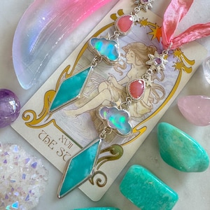 May include: Two silver necklaces with turquoise and opal pendants hang on pink ribbons. The necklaces are displayed with a tarot card, crystals, and polished stones in shades of pink, green, and purple. A pink crescent moon is also present.